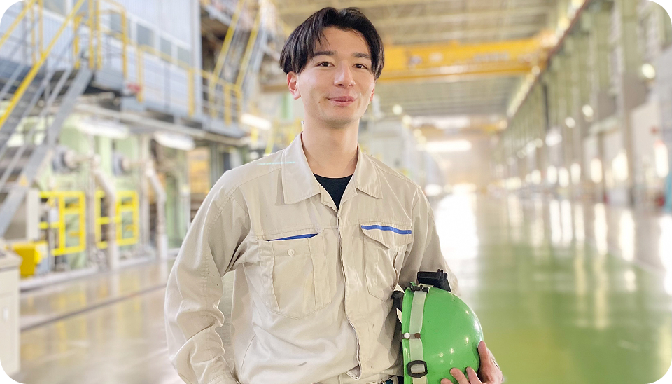 ヘルメットを持ち、作業着を着た男性が工場の中で立っている写真。